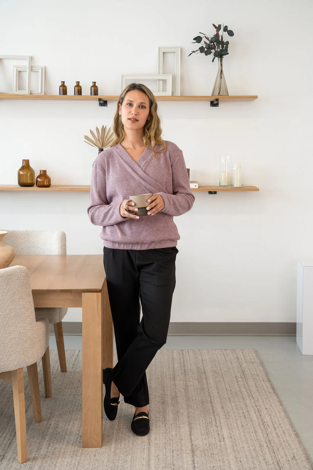 A woman wearing the Mimosa Sweater by Cherry Bobin in Pink, a chunky knit sweater with a faux-wrap neckline, long slightly puffed sleeves, and an elastic waistband. She is wearing it with black pants and holding a mug while standing by a table. 