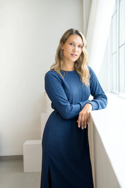 A woman wearing the Negroni Dress by Cherry Bobin in Blue, a mid-calf dress with in cable-knit textured fabric, side slits, and long, slighty puffed sleeves. She is standing near a window in a white room. 