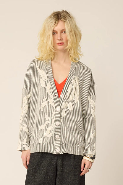 Woman wearing the Gus Cardigan from Cokluch in Natural, with ribbed fabric and floral embellishments on the front and sleeves. It has a relaxed it and five buttons. She is wearing it over an  orange top with grey pants and standing in front of a white background.