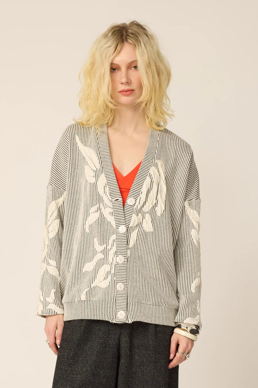 Woman wearing the Gus Cardigan from Cokluch in Natural, with ribbed fabric and floral embellishments on the front and sleeves. It has a relaxed it and five buttons. She is wearing it over an  orange top with grey pants and standing in front of a white background.