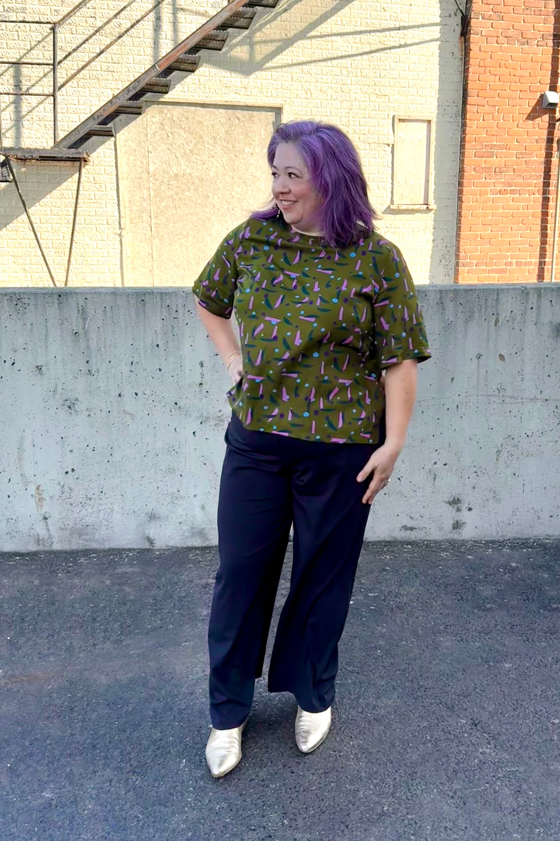 A woman wearing the The Cecil Box Blouse by Mandala in Playtime Ivy print. It has a high neckline, wide 1/2 sleeves, and a side hem slit detail. She is wearing it with black pants and standing outside by a brick wall. 