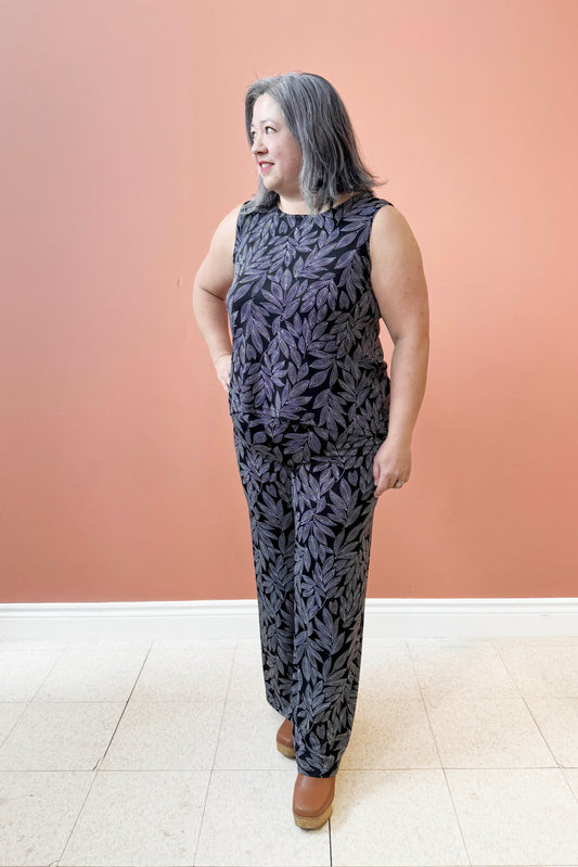 A woman wearing the Swing Cami by Rien ne se Perd in Black and White Foliage, a trapeze cut tank with a hi-low rounded hemline. She is wearing it with the matching Rumba Palazzo pants and standing  in front of a pink wall. 