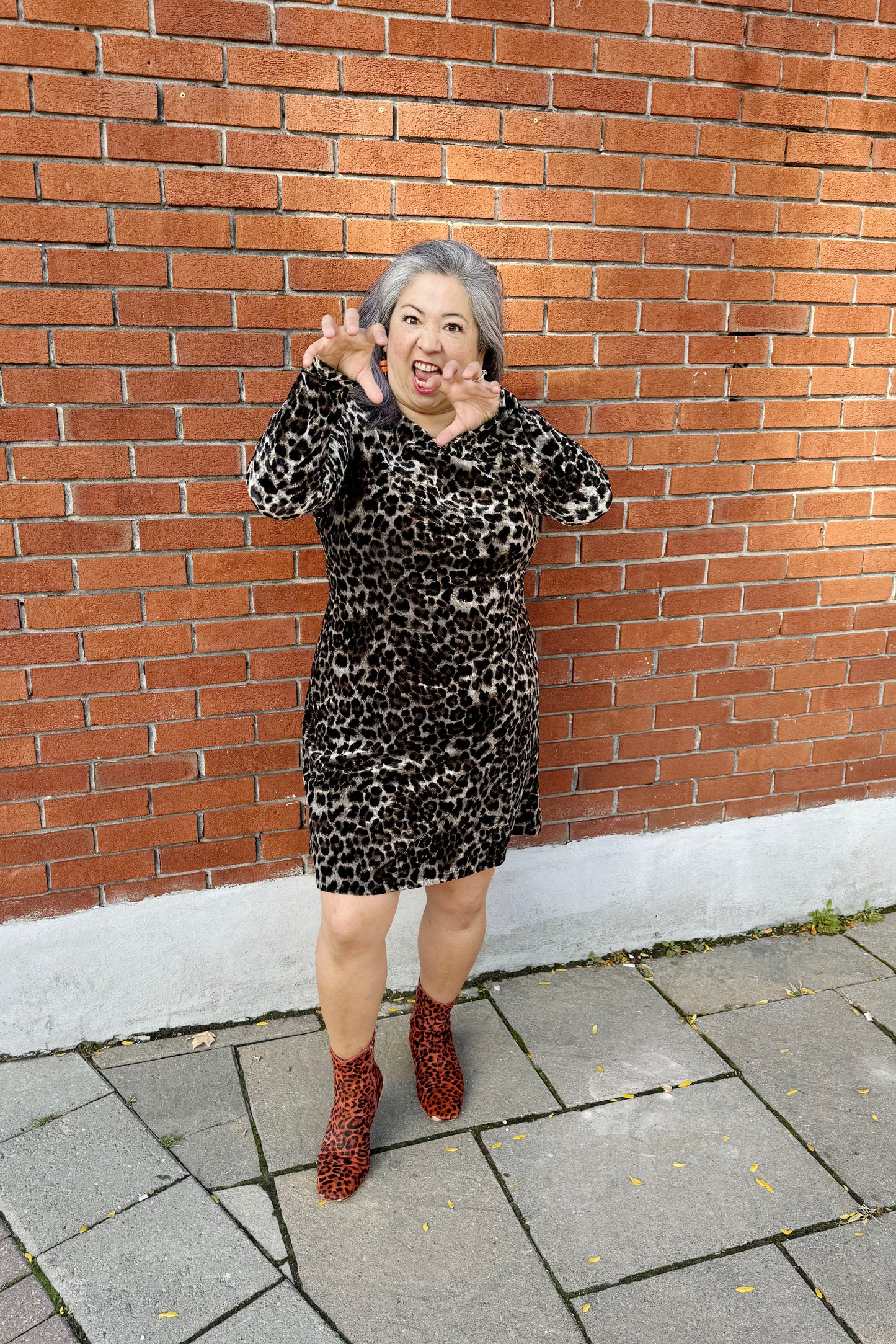 A woman wearing the Luc Fontaine Panthera Dress in Leopard Print, featuring a faux-wrap neckline, long sleeves, fuzzy animal print fabric, a sheath shape, and an above the knee length, standing in front of a brick wall. 
