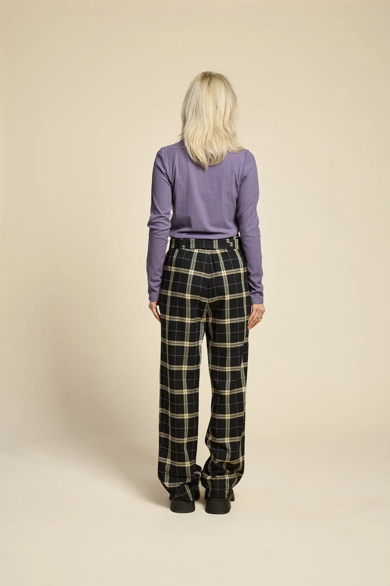 Back view of a woman wearing the Solo Top by Cokluch in Heron, a fitted turtleneck with a tone on tone striped, textured fabric and a twist detail at the neck. She is wearing it with the West Coast pants in Black  Plaid and standing in front of a beige background. 
