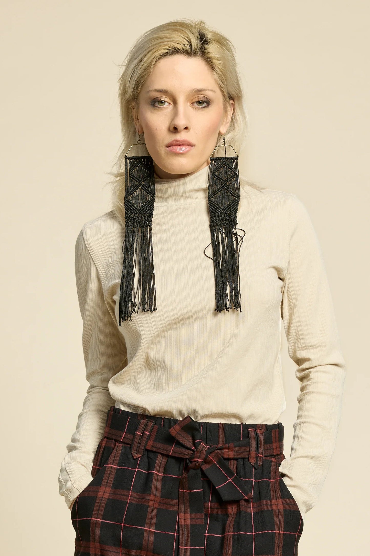 A woman wearing the Solo Top by Cokluch in Sand, a fitted turtleneck with a tone on tone striped, textured fabric and a twist detail at the neck. She is wearing it with the West Coast pants in Burgundy Plaid and standing in front of a beige background. 