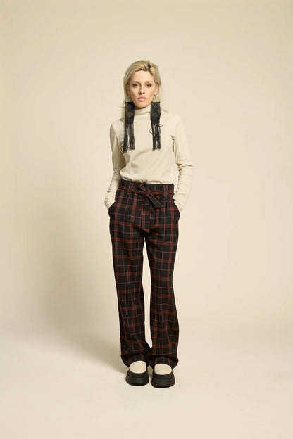 A woman wearing the Solo Top by Cokluch in Sand, a fitted turtleneck with a tone on tone striped, textured fabric and a twist detail at the neck. She is wearing it with the West Coast pants in Burgundy Plaid and standing in front of a beige background. 