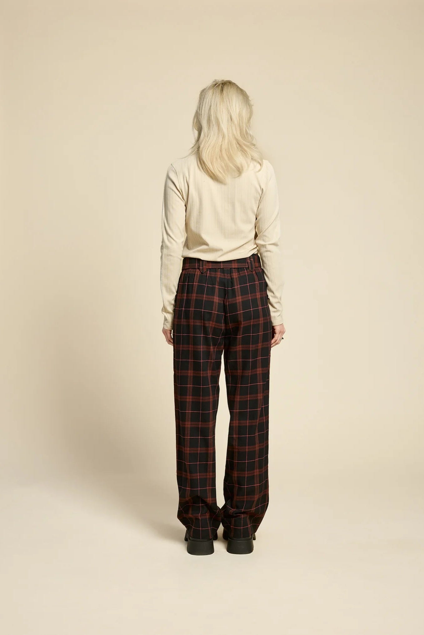 Back view of a woman wearing the Solo Top by Cokluch in Sand, a fitted turtleneck with a tone on tone striped, textured fabric and a twist detail at the neck. She is wearing it with the West Coast pants in Burgundy Plaid and standing in front of a beige background. 