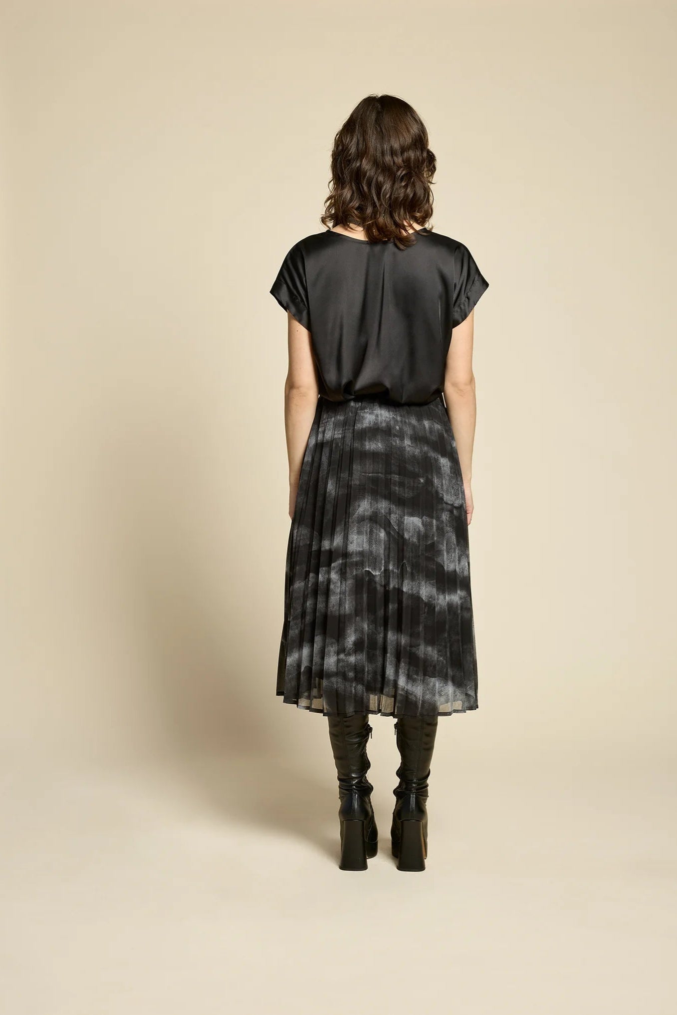 Back view of a woman wearing the Flowers Skirt by Cokluch in Mist Black, a midi-length pleated skirt with a sheer overlayer and an elastic waist. She is wearing it with a black top and standing in front of a beige background. 