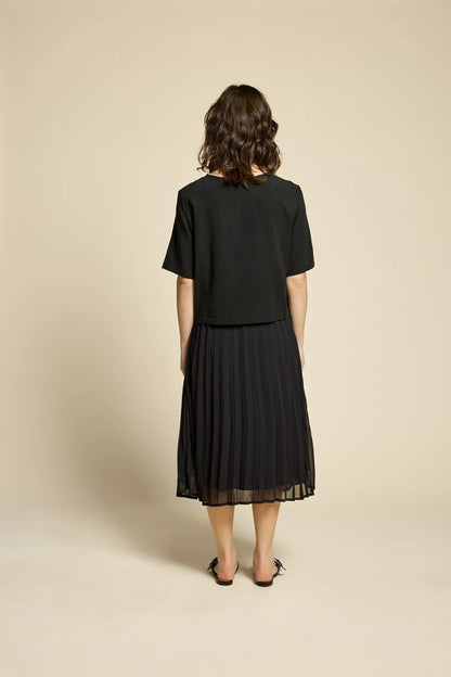 Back view of a woman wearing the Flowers Skirt by Cokluch in Black, a midi-length pleated skirt with a sheer overlayer and an elastic waist. She is wearing it with a black top and standing in front of a beige background. 
