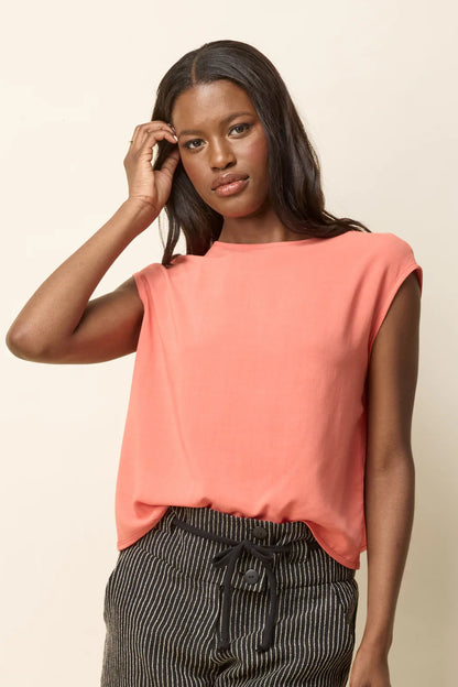 A woman wearing the Bayang Shirt by Cokluch in Papaya, featuring a  round neck, short extended sleeves, and a cross-over bottom hem at the back. She is wearing it with striped shorts and is standing in front of a beige background. 