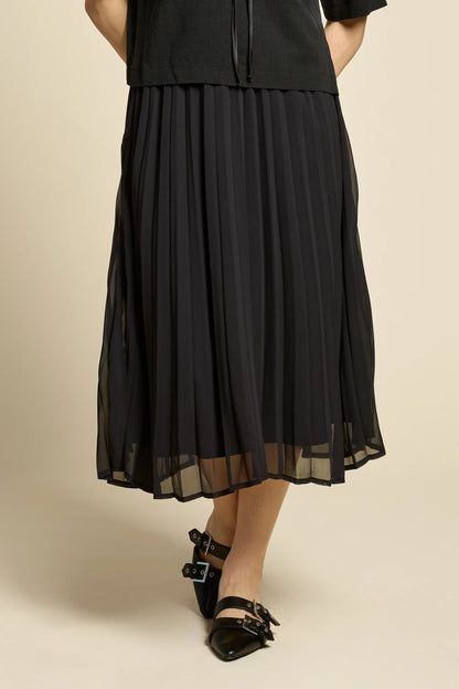 Waist down view of a woman wearing the Flowers Skirt by Cokluch in Black, a midi-length pleated skirt with a sheer overlayer and an elastic waist. She is wearing it with a black top and standing in front of a beige background. 