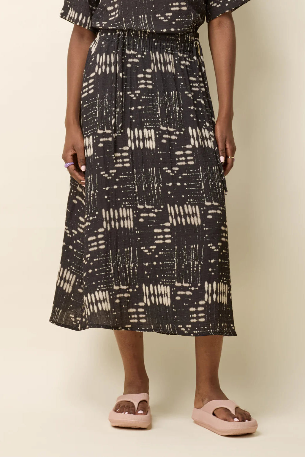 Waist-down view of a woman wearing the Susan Skirt by Cokluch in Black Ink, an ankle-length skirt in a geometric print with an elastic waist with double drawstrings, a side slit, and side pockets. She is standing in front of a beige background. 