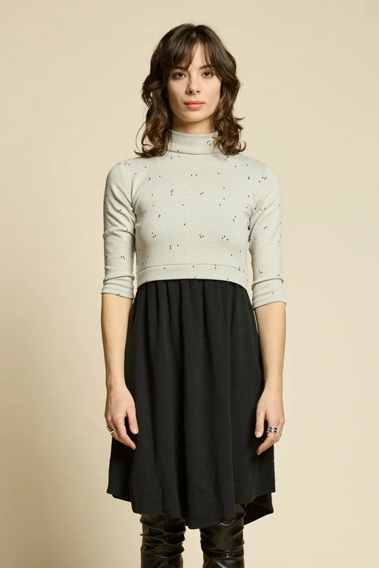 A woman wearing the Crystal Dress by Cokluch, featuring a knit top half in Natural with a flowing black skirt that falls to the knee. It has a high neck, 3/4 sleeves, and an opening at the back. She is standing in front of a beige background. 
