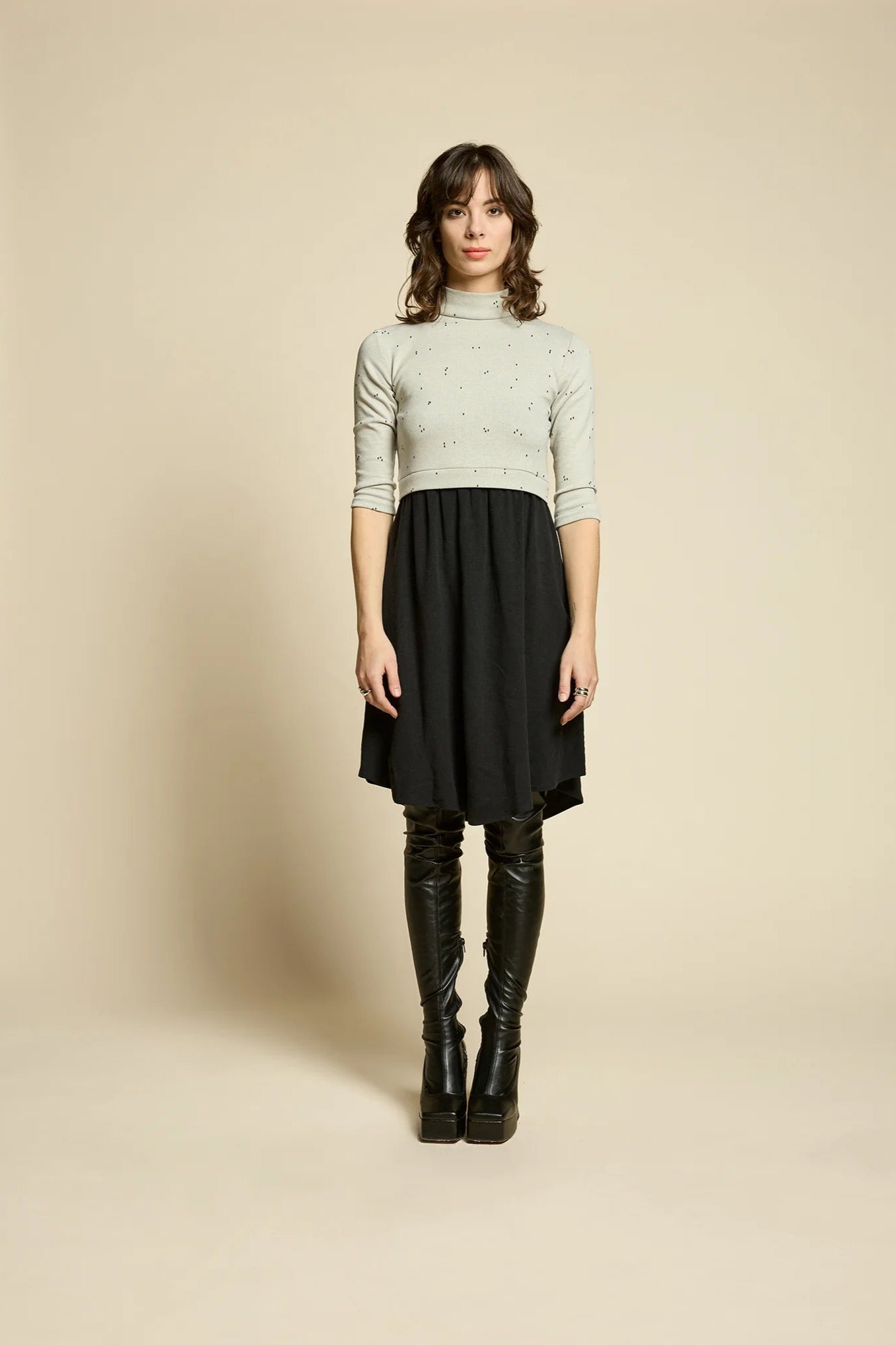 A woman wearing the Crystal Dress by Cokluch, featuring a knit top half in Natural with a flowing black skirt that falls to the knee. It has a high neck, 3/4 sleeves, and an opening at the back. She is standing in front of a beige background. 