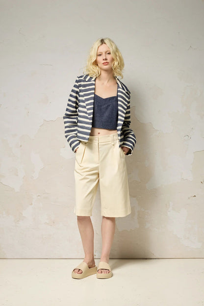 A woman wearing the Vesta Jacket by Cokluch in Denim Stripe over a denim top and beige shorts against a plain background