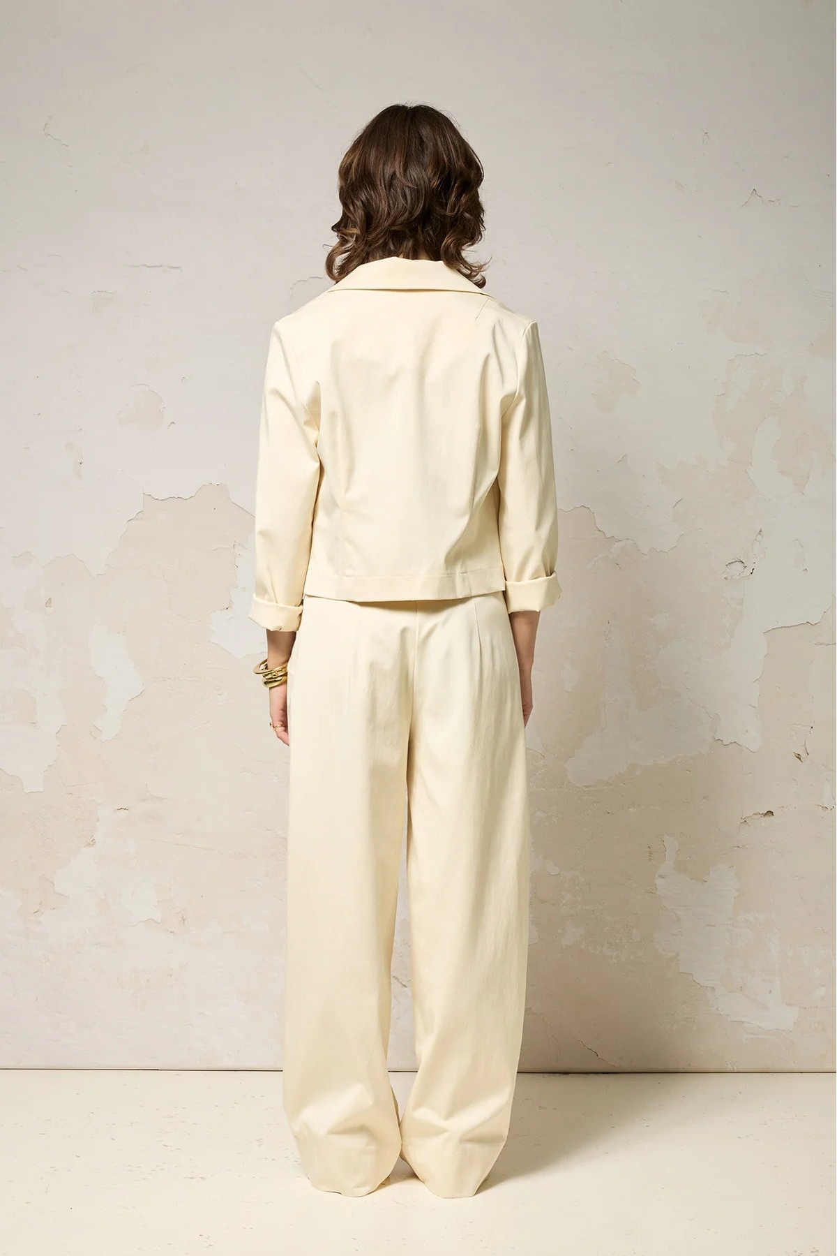 Back view of a woman wearing the Vesta Jacket by Cokluch in Vanilla over a patterned top and matching pants against a plain background