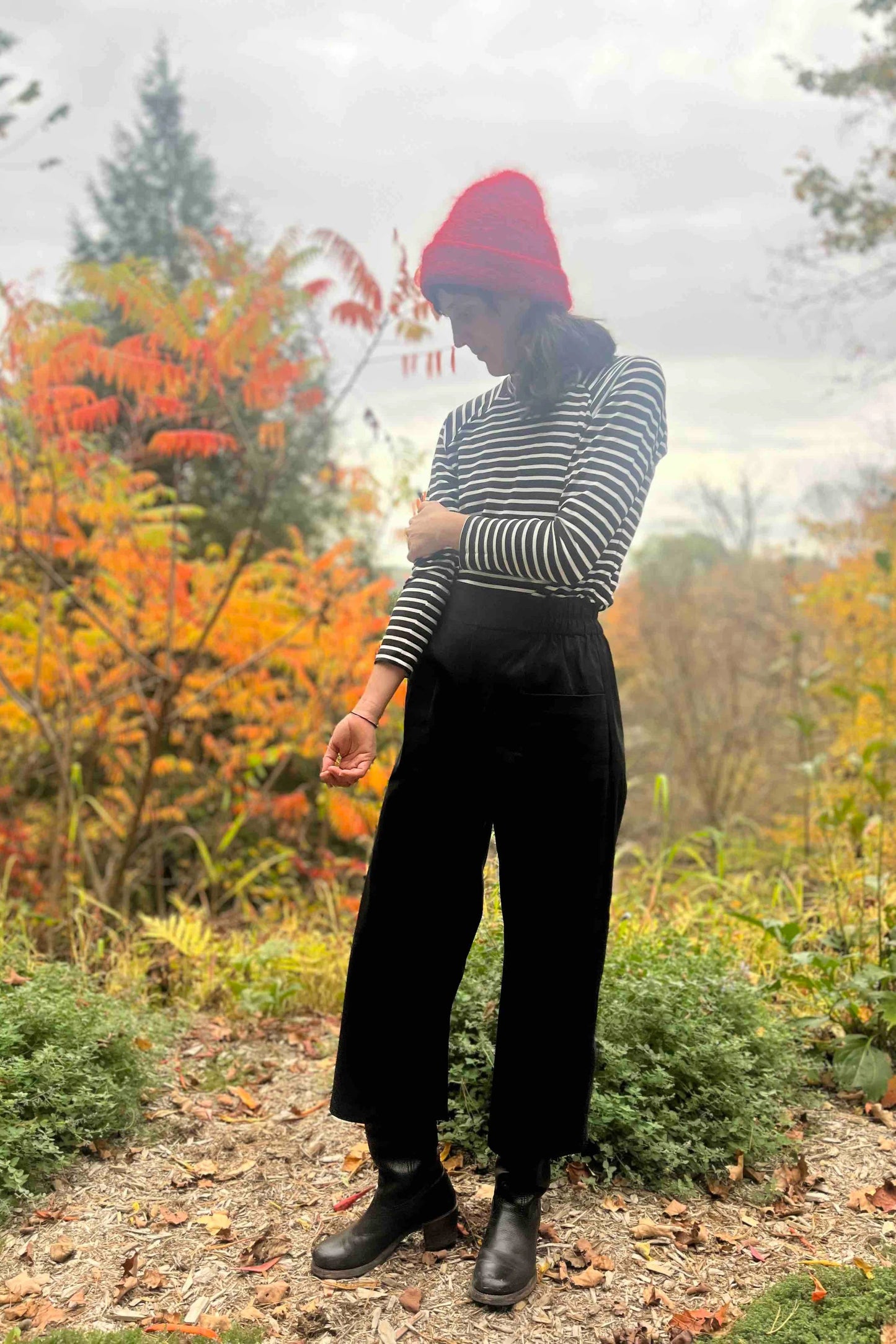 A woman wearing the Olea Pants by 
Kazak in Black, wide-legged ankle-length pants with a flat-front elastic waist and large patch pockets. She is wearing them with the Oris Turtleneck in Black Stripe and standing in a forest.