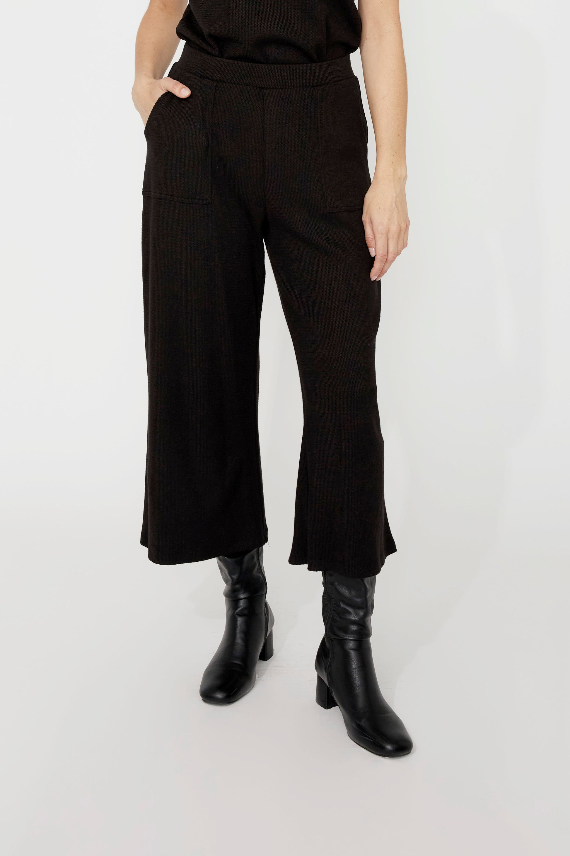 Wait down view of a woman wearing the Penelope Wide Leg Knit Pants by Compli K in Black, cropped wide leg pants in thick textured knit with large patch opckets and an elastic waist. She is wearing them a black top and standing in front of a grey background. 