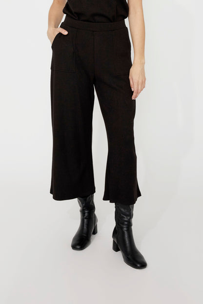 Wait down view of a woman wearing the Penelope Wide Leg Knit Pants by Compli K in Black, cropped wide leg pants in thick textured knit with large patch opckets and an elastic waist. She is wearing them a black top and standing in front of a grey background. 