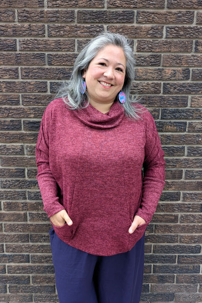 A woman wearing the Nash Sweater by Compli K in Garnet, with a cowl neck, long extended sleeves, angled vertical seams on the front, and front patch pockets. She is standing outside in front of a brick wall.