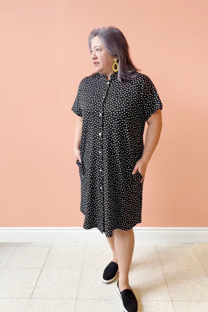 Christina is wearing the Cora Dress by Pure Essence in Black with polka dots, standing in front of a coral wall
