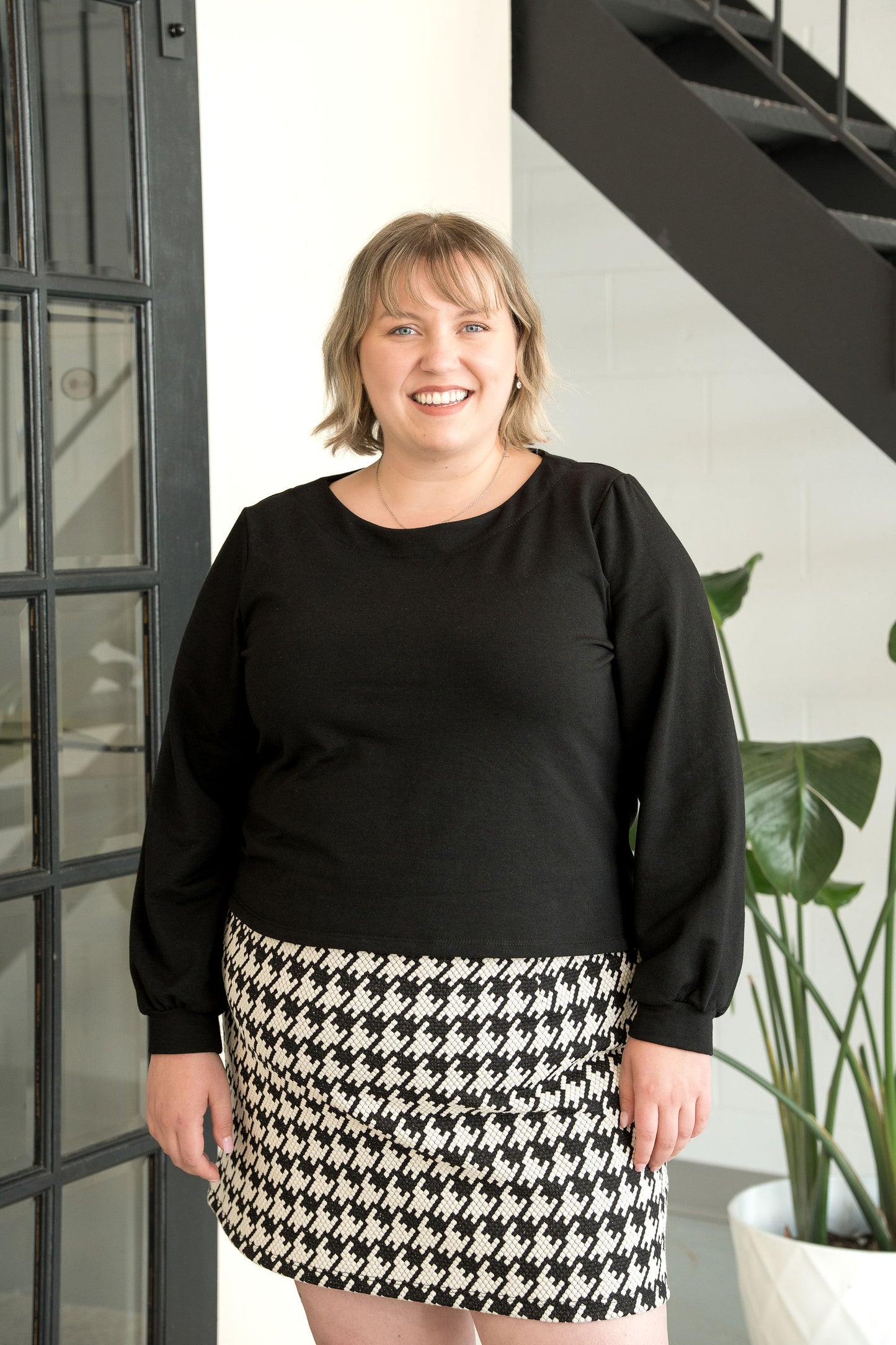 A woman wearing the Cortado Skirt by Cherry Bobin in Houndstooth, a short fitted skirt with an elastic waistband and invisible side zipper. She is wearing it with a black top and standing in front of a plant. 