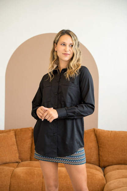 A woman wearing the Cortado Skirt by Cherry Bobin in Jaquard Zigzag, a short fitted skirt with an elastic waistband and invisible side zipper. She is wearing it with a black top and standing in front of a couch.