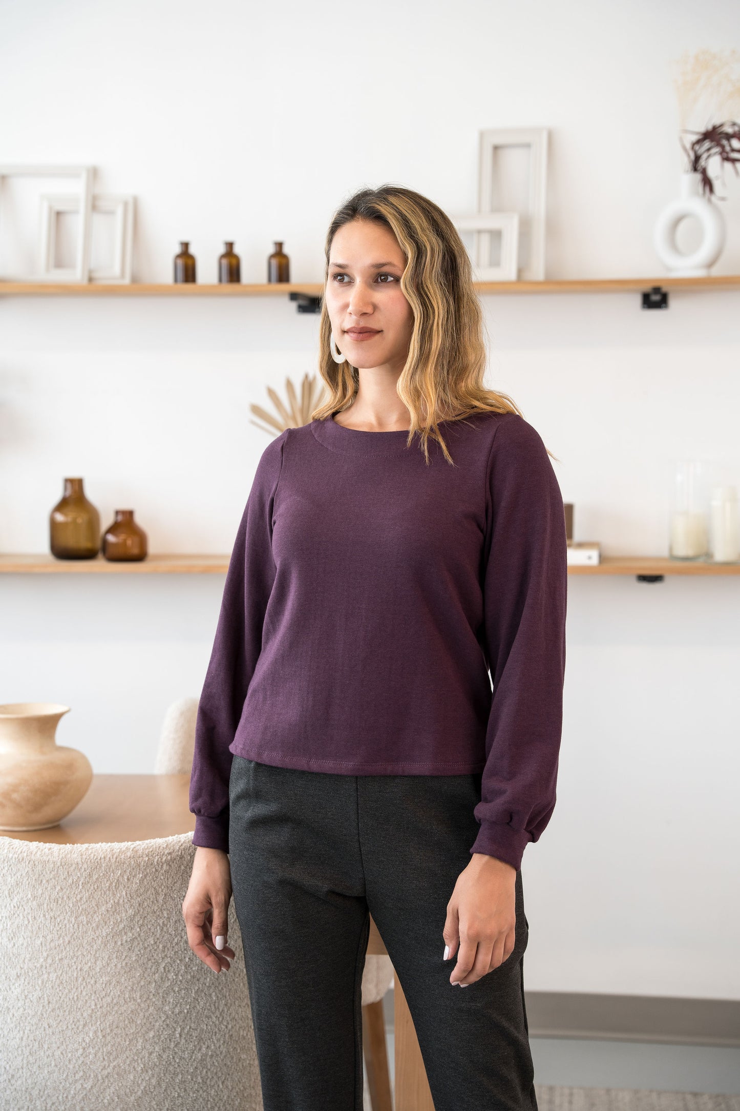 A woman wearing the Cosmopolitain Sweater by Cherry Bobin in Mauve, with a rounded neck and long sleeves with a puff detail. She is wearing it with black pants and standing in a room with shelves. 
