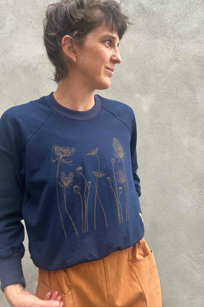 A woman wearing the Marilou Crewneck with Flowers by Kazak in Navy, a raglan sleeved sweatshirt with a screen print design. She is wearing it with yellow and standing by a grey wall.