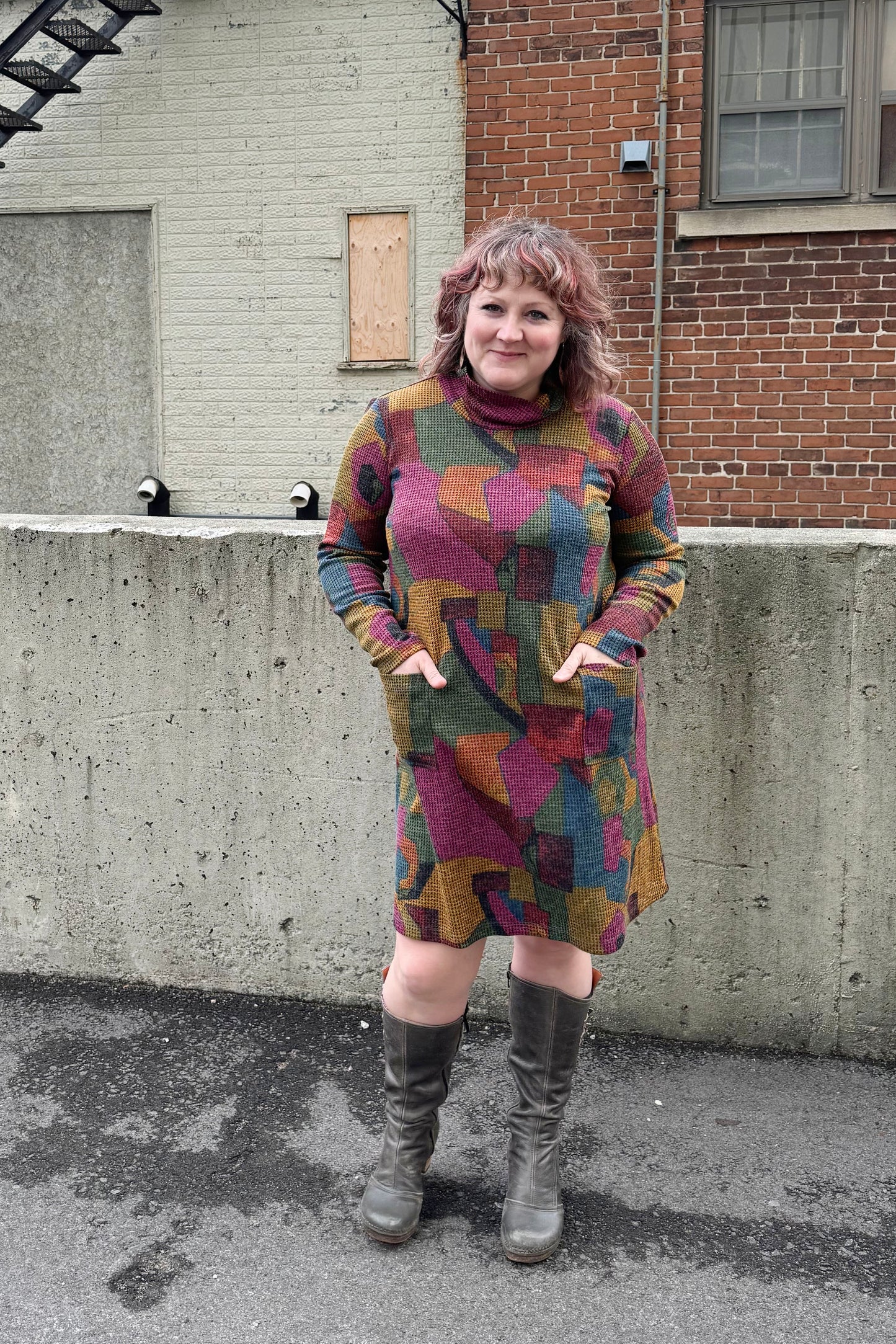 A woman wearing the Damon Dress by Pure Essence in Berry/Mustard, featuring a cowl neck, long sleeves, patch pockets, and a knee length hem. She's standing outside in front of a brick building. 