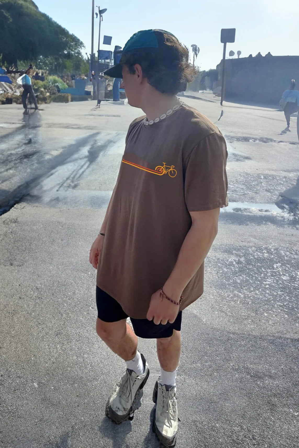 A man wearing the Disco Bike T-shirt by Message Factory in Nutmeg, featuring a retro-inspired cycling logo in orange and yellow. He is wearing it with black shorts and standing outside. 