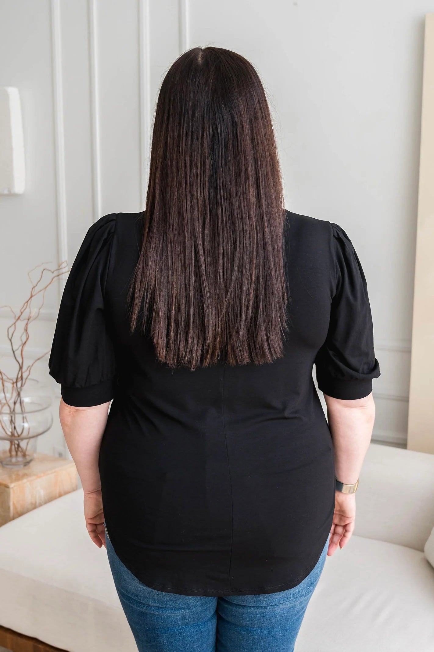 A woman wearing the Darling Tee by Dotty in Black, featuring a v-neck, pleated shoulders, and elbow length sleeves, with jeans. She is standing n front of a white sofa in a room with white walls 