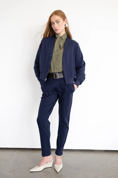 A woman wearing the Douglas Fleece Cardigan by Eve Lavoie in Navy, a short cardigan with navy buttons and a wide waistband. She is wearing it with the Blair Shirt  in Olive nd the Fraser Pants in Navy and standing against a white background