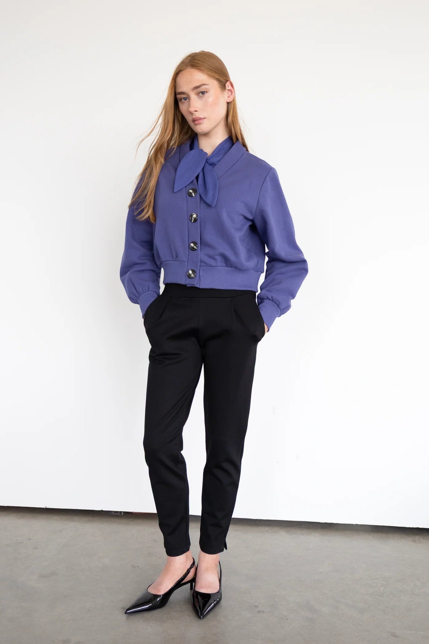 A woman wearing the Douglas Fleece Cardigan by Eve Lavoie in Purple, a short cardigan with tortoiseshell buttons and a wide waistband. She is wearing it with the Blair Shirt  in Purple and the Fraser Pants in Black and standing against a white background