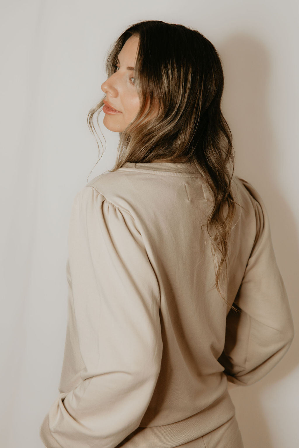 Back waist up view of a woman wearing the Tasia Sweatshirt by Betty x Bow in Crema, an oversized crew neck with subtly puffed shoulders, ribbing a the cuffs, hem and collar, and a monochromatic embroidered bow logo. She is standing in front of a white background. 