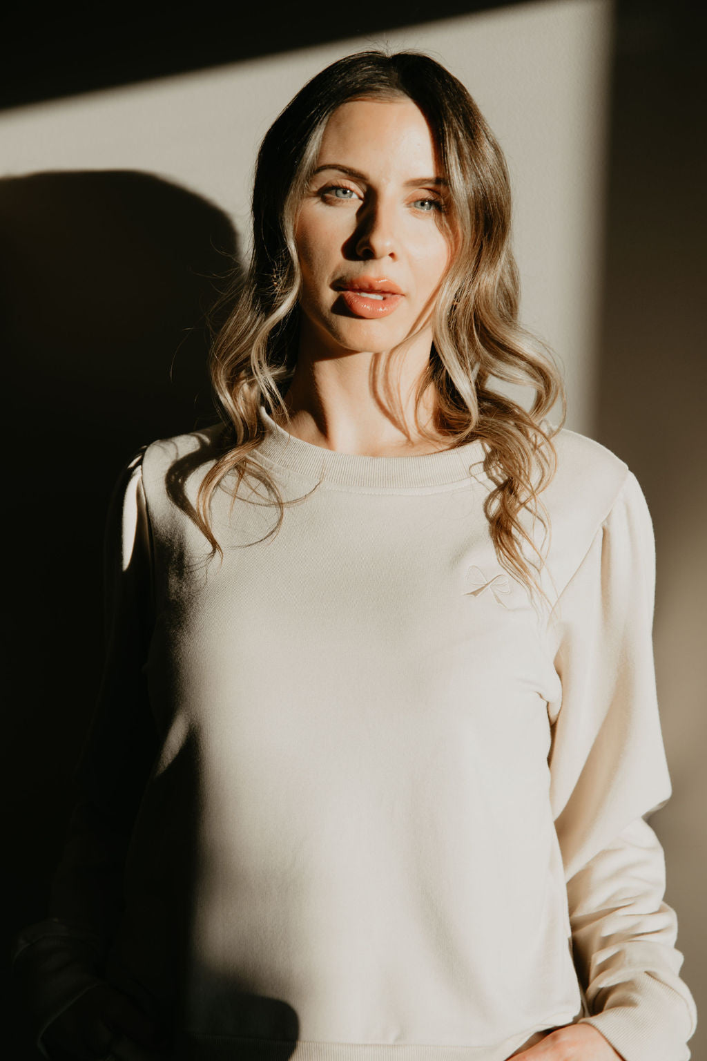 Waist up view of a woman wearing the Tasia Sweatshirt by Betty x Bow in Crema, an oversized crew neck with subtly puffed shoulders, ribbing at the cuffs, hem and collar, and a monochromatic embroidered bow logo. 