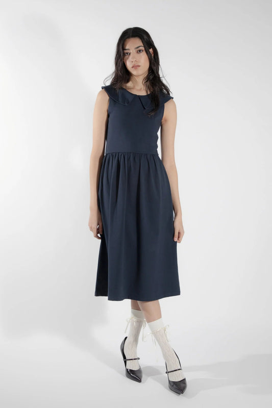 A woman wearing the Lobelie Reversible Sun Dress by Eve Lavoie in  Navy, a sleeveless dress with a shawl collar on one side and a Claudine collar on the other, a snug bodice and full gathered skirt, and below the knee hem. She is standing in front of a white background. 