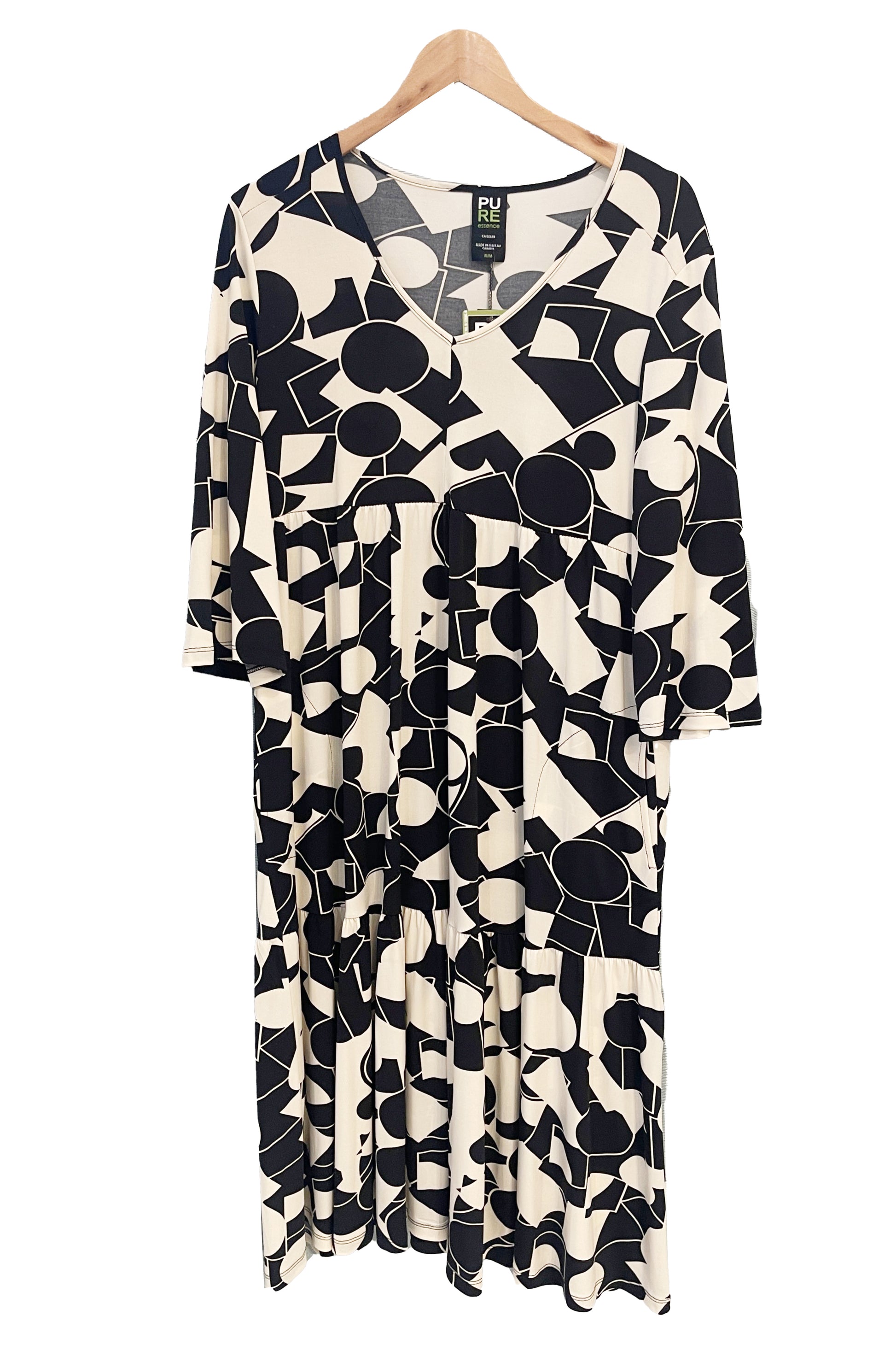 The Farah Dress by Pure Essence in Black/Ivory, a geometric print dress with a v-neck, 3/4-sleeves, baby doll shape, tiered midi length skirt, and front patch pockets, is shown hanging front of a white background. 