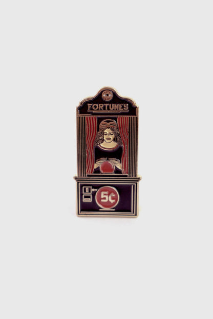 The Fortune Teller Pin from Badaboöm Studio is an enamel pin shaped like a vintage fortune telling booth with the word 'FORTUNES' at the top and '5c' on the lower panel. It's shown against a white background. 