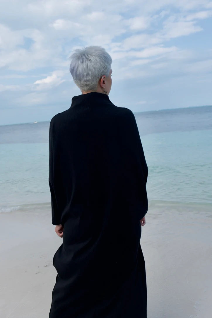 Back view of a woman wearing the Frankfurt Dress by MAS in Pepper, a One Size ribbed cotton dress with a mock turtleneck, long batwing sleeves, and midi length. She is standing on a beach.