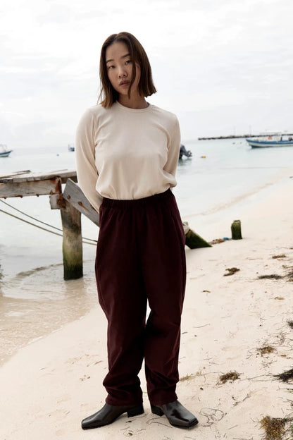 A woman wearing the Funchal Pants by MAS in Fig, fine wale corduroy pants with an elastic waist and straight cut legs. She is wearing them with a white top and is standing on a beach. 
