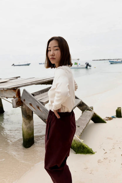 Sid view of a woman wearing the Whistler Long Sleeve Top in Almond, a ribbed cotton knit with a round neck and straight cut. She is wearing it with the Funchal Pants in Fig and standing on a beach. 