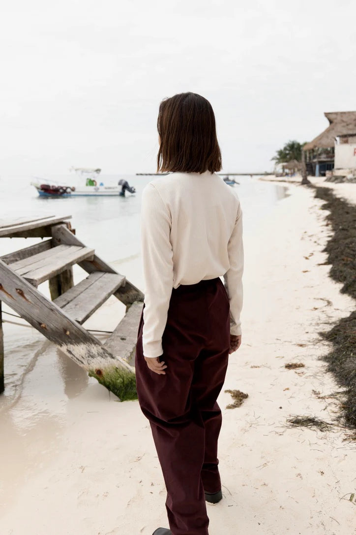 Back view A woman wearing the Whistler Long Sleeve Top in Almond, a ribbed cotton knit with a round neck and straight cut. She is wearing it with the Funchal Pants in Fig and standing on a beach. 
