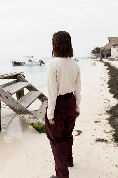 Back view A woman wearing the Whistler Long Sleeve Top in Almond, a ribbed cotton knit with a round neck and straight cut. She is wearing it with the Funchal Pants in Fig and standing on a beach. 
