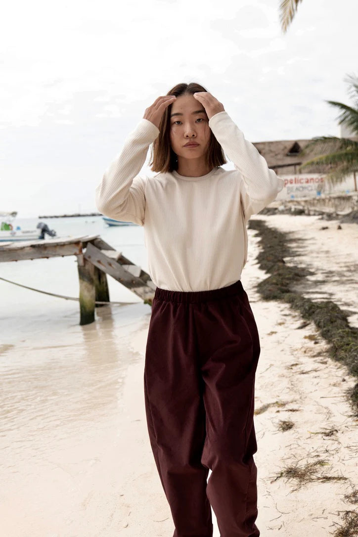 A woman wearing the Funchal Pants by MAS in Fig, fine wale corduroy pants with an elastic waist and straight cut legs. She is wearing them with the Whistler top in Almond and is standing on a beach. 