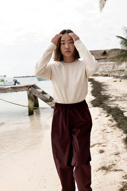 A woman wearing the Funchal Pants by MAS in Fig, fine wale corduroy pants with an elastic waist and straight cut legs. She is wearing them with the Whistler top in Almond and is standing on a beach. 