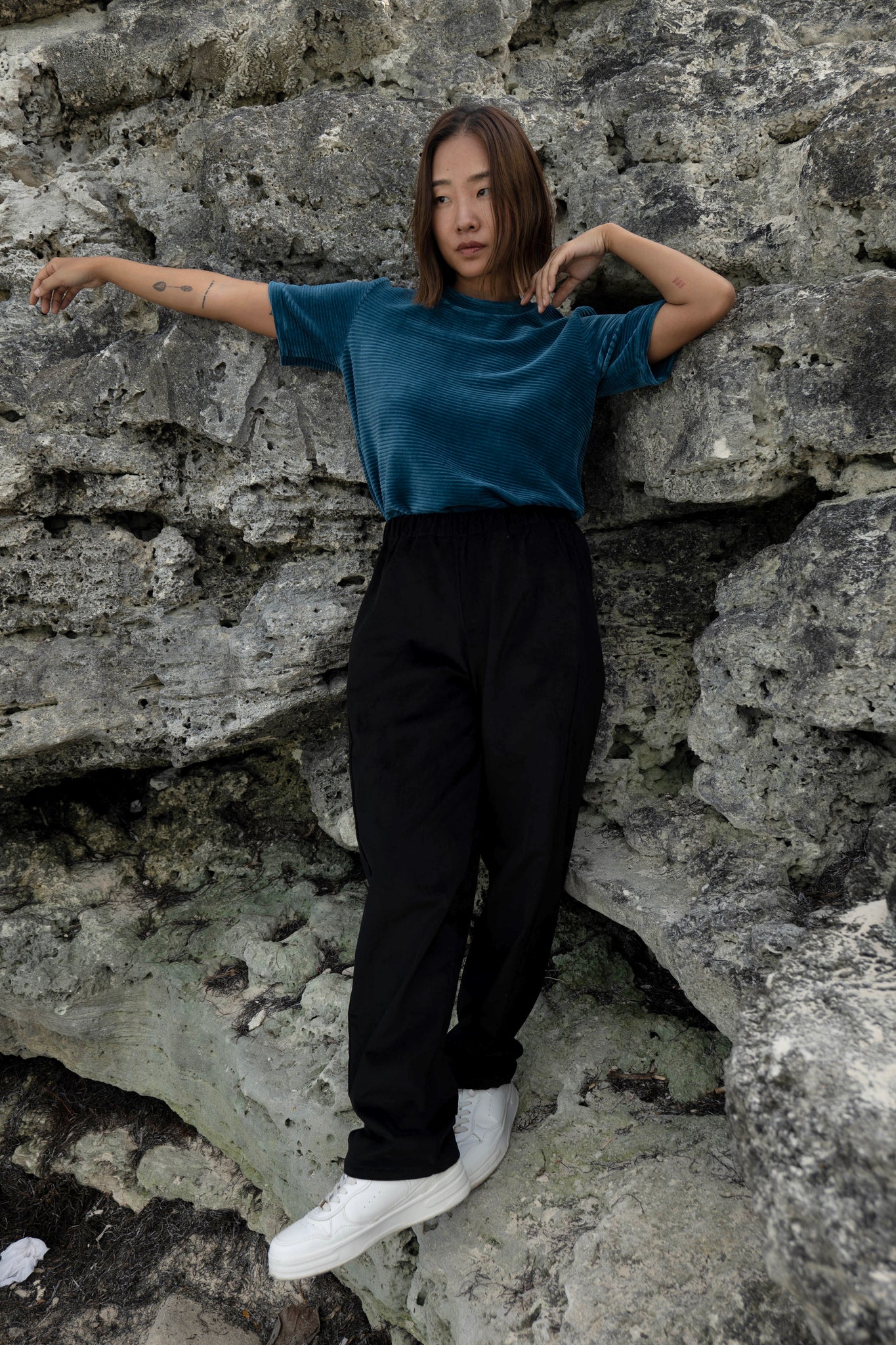 A woman wearing the Tofino T-Shirt by MAS in Juniper, a short sleeved top in ribbed velvet. She is wearing it with the Funchal Pants in Pepper and standing against a rock wall. 