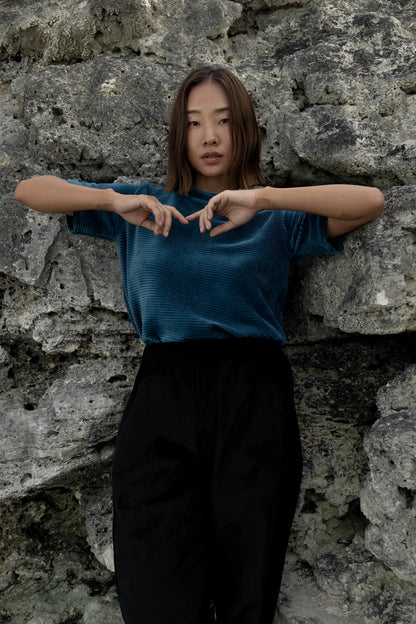 A woman wearing the Tofino T-Shirt by MAS in Juniper, a short sleeved top in ribbed velvet. She is wearing it with the Funchal Pants in Pepper and standing against a rock wall. 