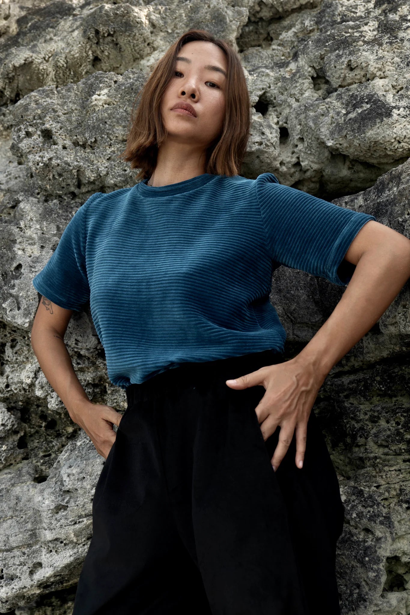 A woman wearing the Tofino T-Shirt by MAS in Juniper, a short sleeved top in ribbed velvet. She is standing in front of a rock wall. 