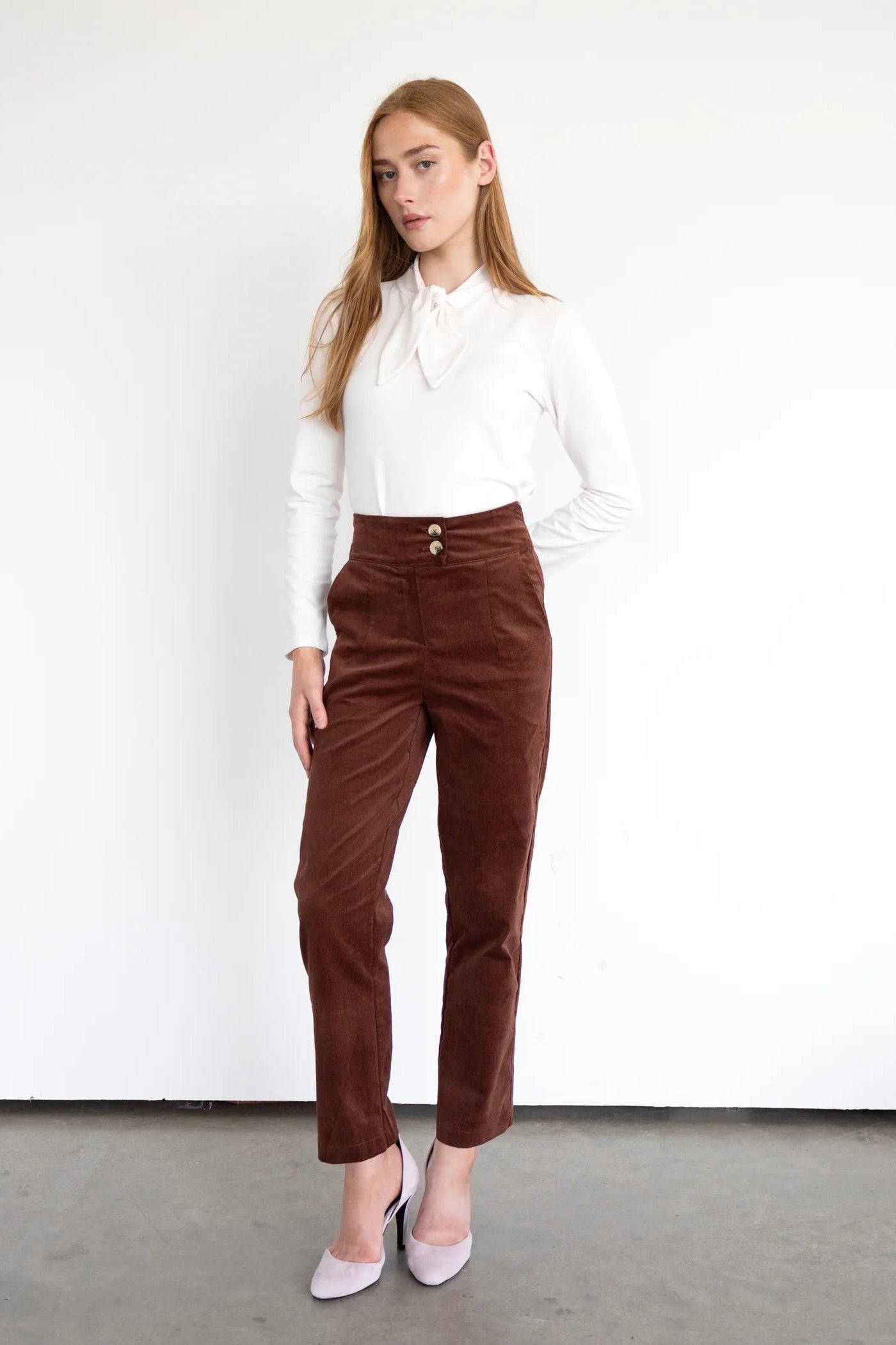 A woman wearing the Graham Corduroy Pants by Eve Lavoie in Hazelnut, with a high waist, double button, slim fit and ankle length leg. She is wearing them with a white top and standing in front of a white background. 